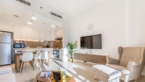A modern kitchen with white cabinets, a dining area with white chairs, a large flat-screen TV mounted on the wall, and a beige sofa in the living area. - Balcony Overlooking the Swimming Pool, Just Minutes from the Park (Dubai)