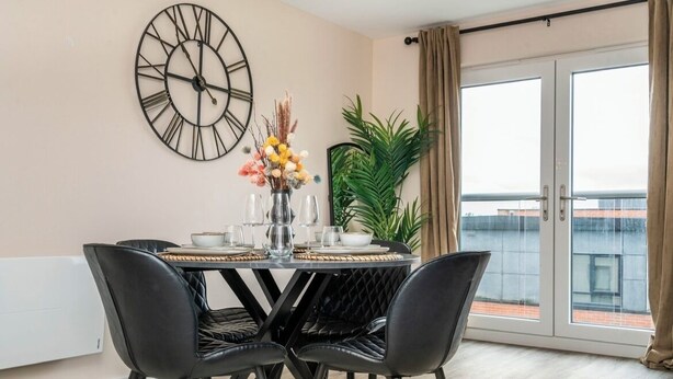 A dining area with a round table set for four, a large wall clock, and a potted plant. - Modern 2bed Manchester+ parking (Manchester)