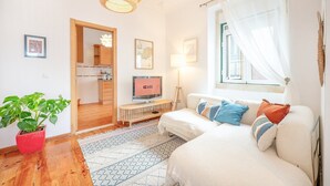 Un salon moderne avec un canapé, un téléviseur, une plante en pot et un coin repas visible à travers une porte ouverte.