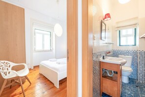 A bedroom with a bed, a white chair, and a window. The bathroom has a sink, mirror, and toilet.