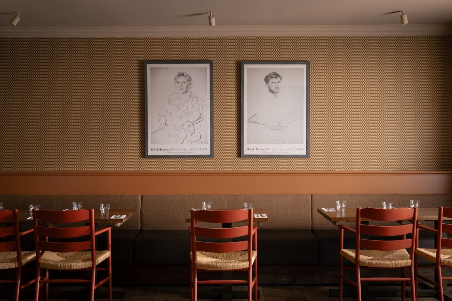 A restaurant interior with framed artwork on the wall, wooden tables, and red chairs.