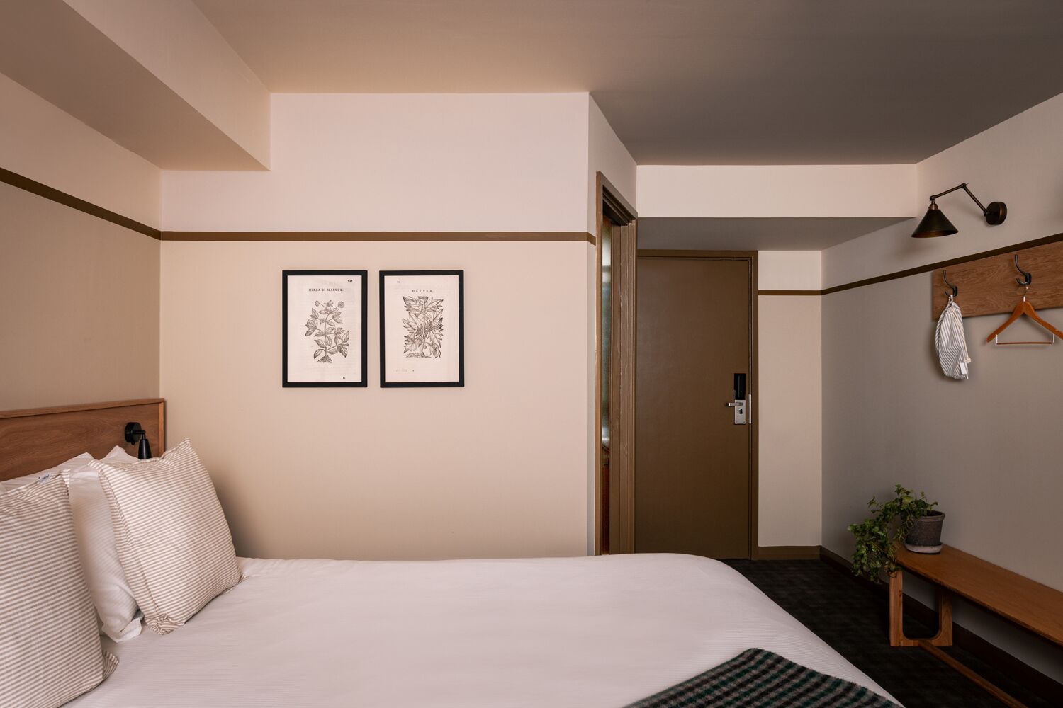 Un dormitorio con una cama, dos láminas botánicas enmarcadas en la pared, una lámpara de noche, una prenda colgada y una planta en maceta.