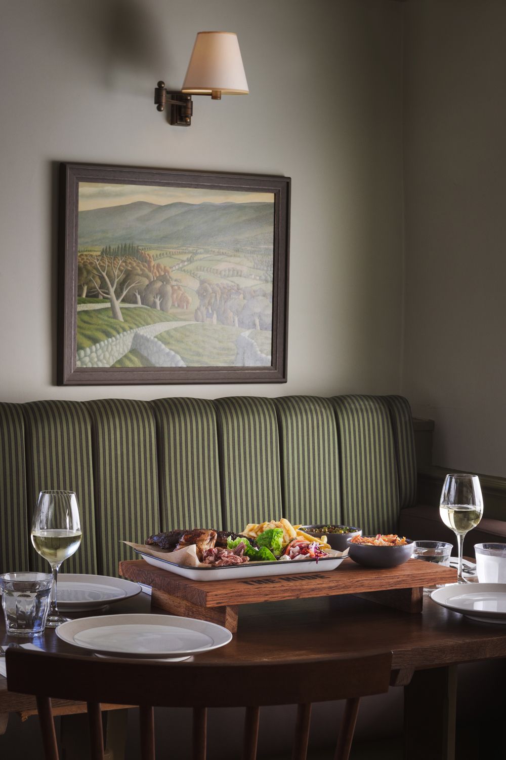 Een eethoek met een houten tafel gedekt voor twee personen, een groene gestoffeerde bank en een lijstwerk schilderij van een landschap aan de muur.
