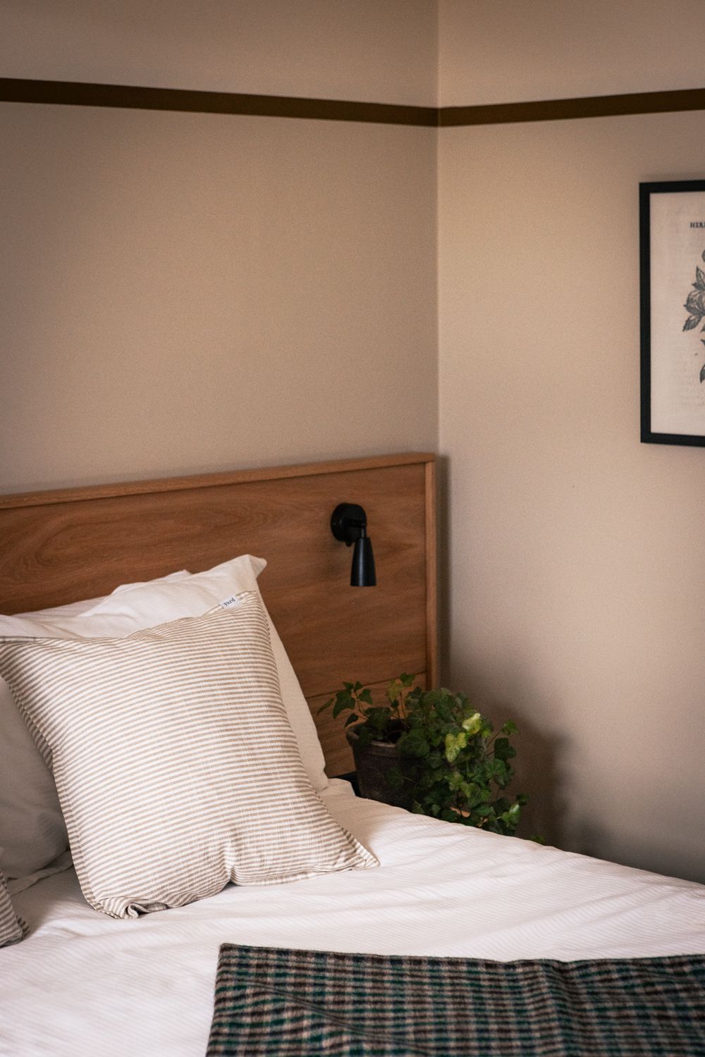 Una cama bien hecha con almohadones a rayas, un cabecero de madera, una lámpara de noche e impresión enmarcadas de motivos botánicos en la pared.