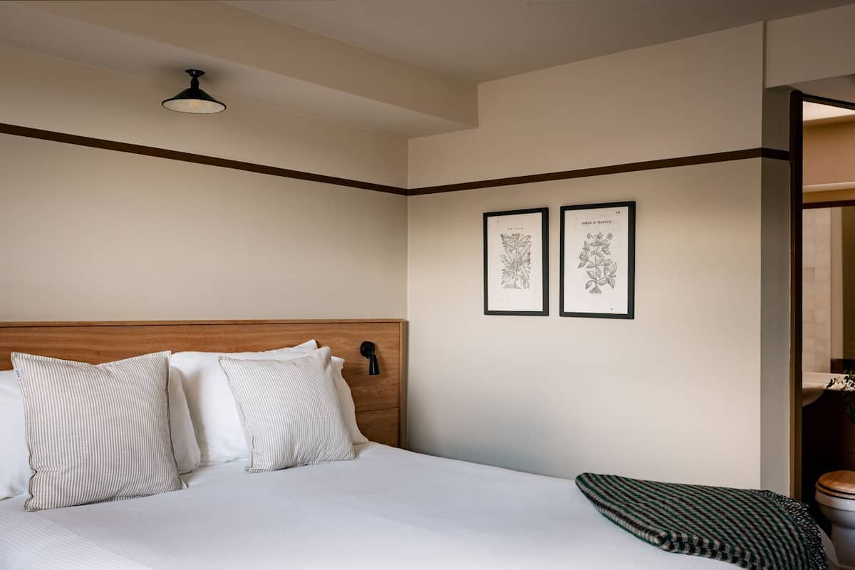 A hotel room with a wooden headboard, a bed with white bedding, two framed botanical illustrations on the wall, and a small bedside table with a lamp.