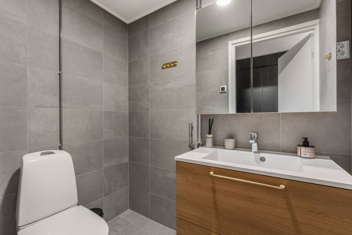 Un baño con baldosas grises, un inodoro blanco, un lavabo con un mueble de madera y un espejo sobre el lavabo.