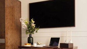 Une chambre d’hôtel avec un bureau en bois, un ordinateur portable, un vase avec des fleurs, un lit recouvert d’une couverture à motifs et un grand écran noir fixé au mur.