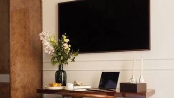 A hotel room with a wooden desk, a laptop, a vase with flowers, a bed with a patterned blanket, and a large black screen on the wall.