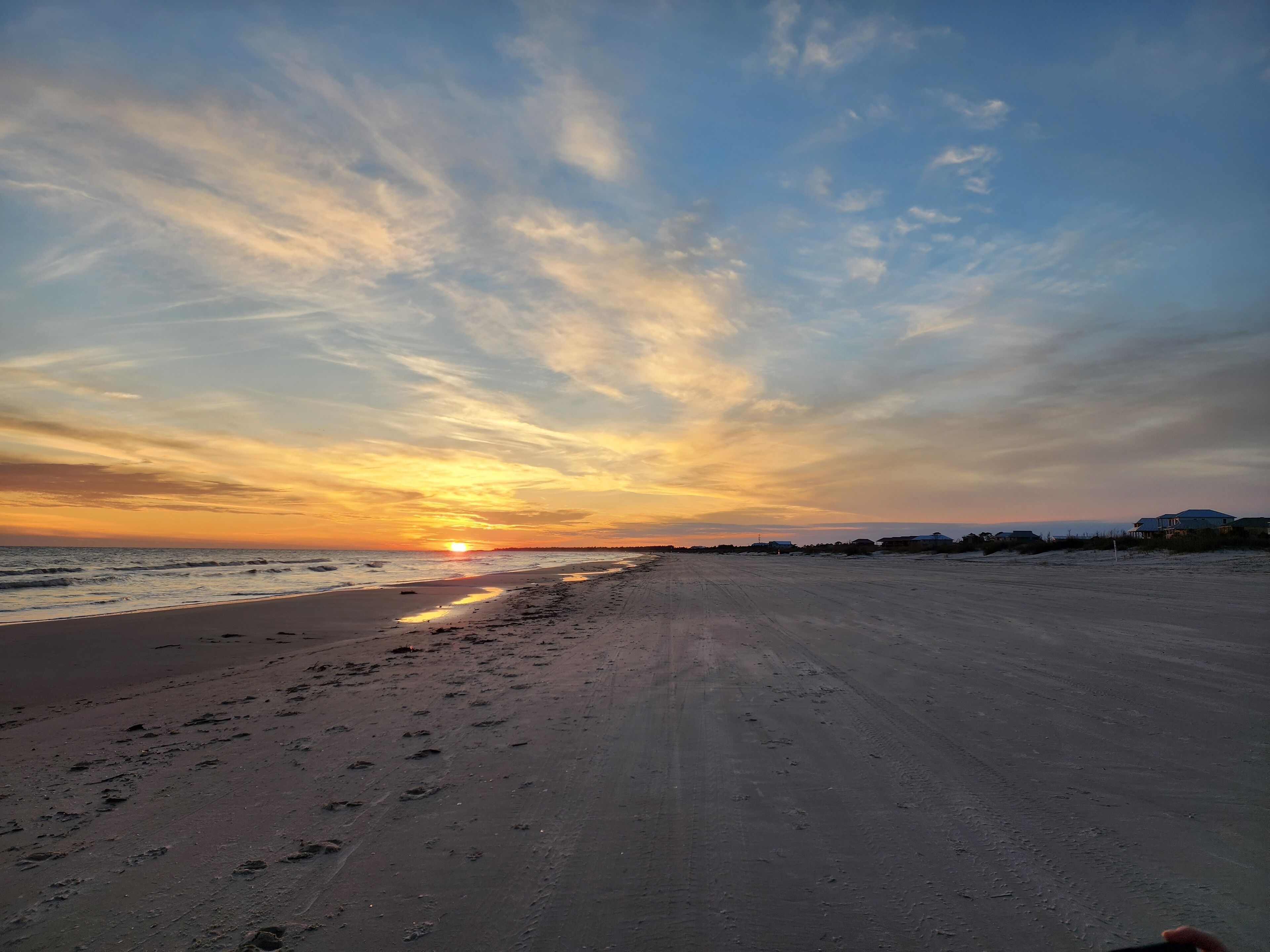 A beach with footprints leading to the water, a sunset in the background, and buildings in the distance.