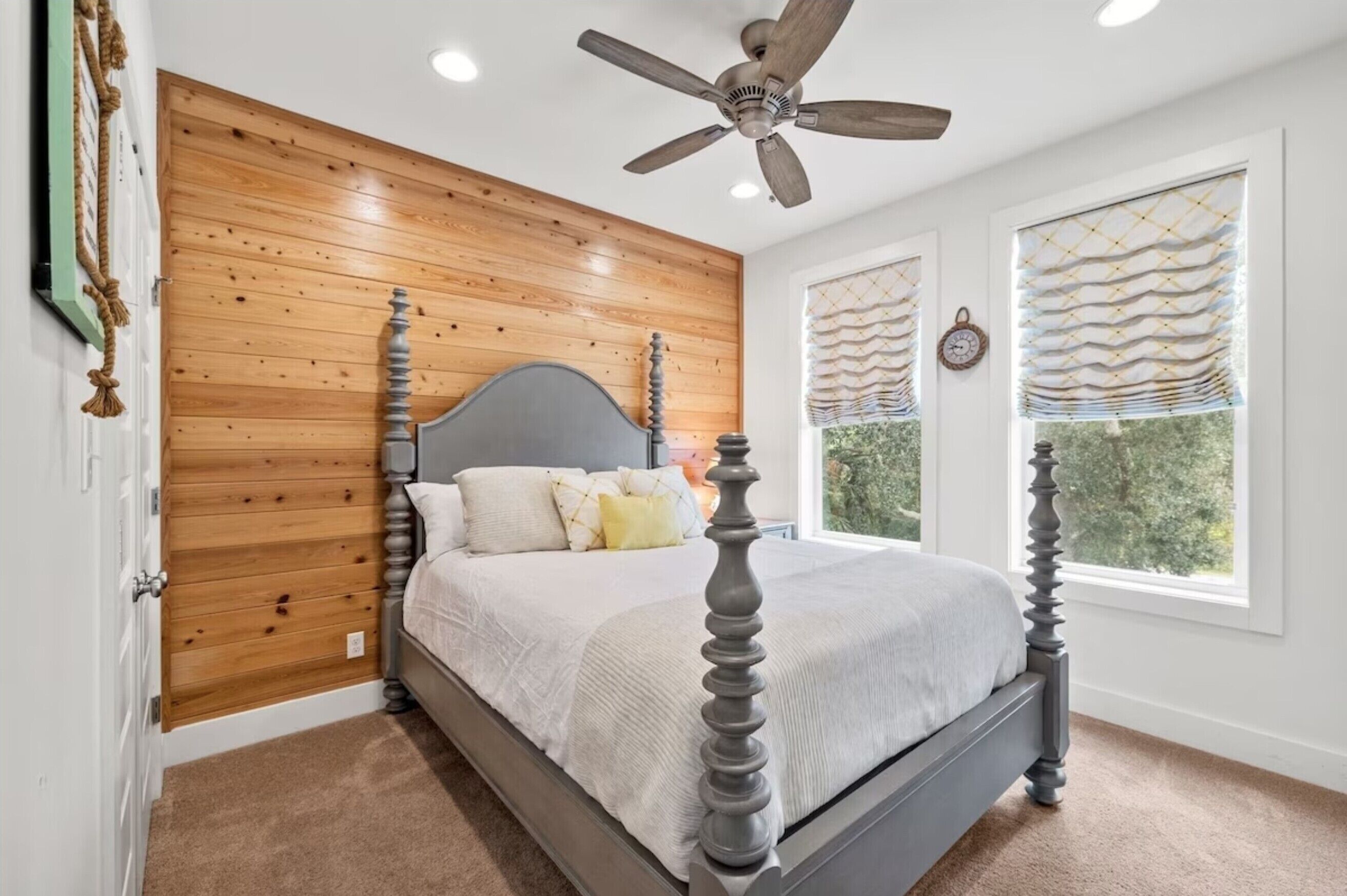 A bedroom with a wooden panel wall, a four-poster bed, a ceiling fan, and large windows with blinds.