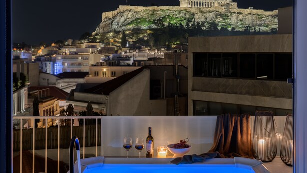 A balcony with a view of a historic building on a hill, a bottle of wine, two glasses, and a bowl of grapes. - Acropolis Penthouse Plunge Pool (Athina)