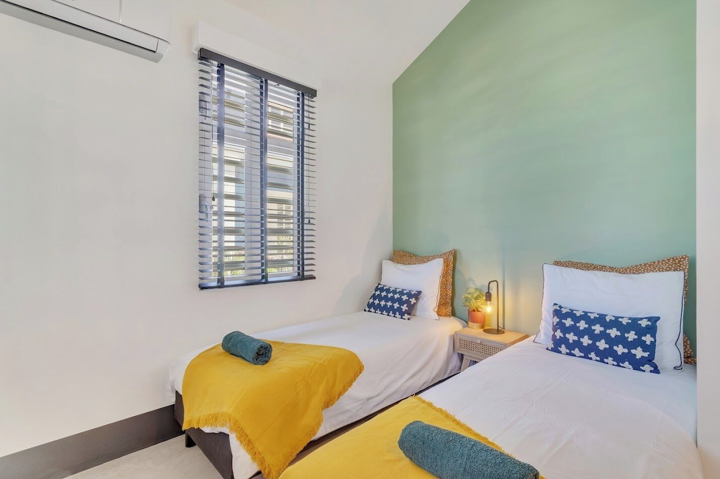 Una habitación con dos camas, una ventana con persianas, una mesita de noche con lámpara y una pared con detalles en verde.