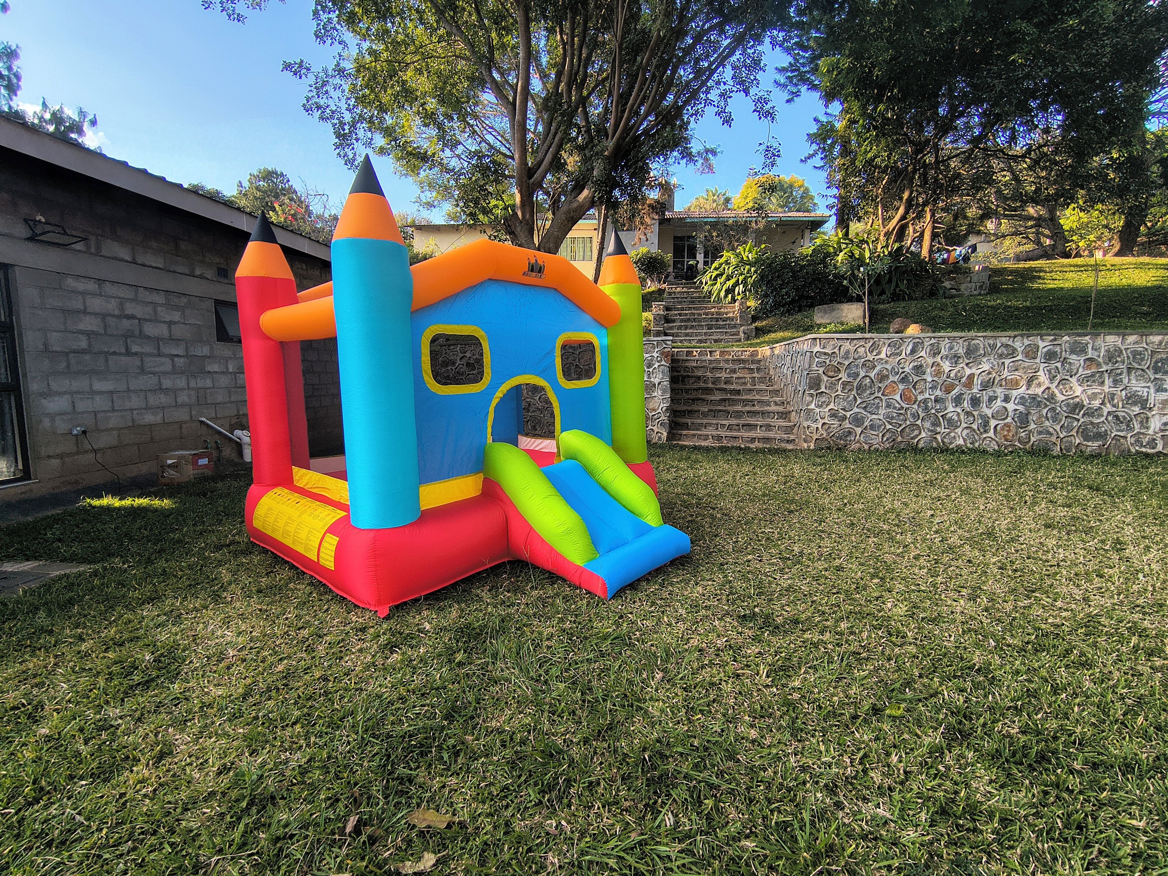 A colorful inflatable play structure in a grassy yard with a stone wall and a house in the background.