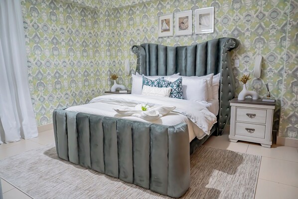 A neatly made bed with a grey upholstered headboard, white bedding, and decorative pillows. There is a bedside table with a vase and a lamp. The room has patterned wallpaper and a light-colored rug. - Amazing & Solemn Ocean & Beach Front Apartment in Juan Dolio with a Huge Balcony (Guayacanes)