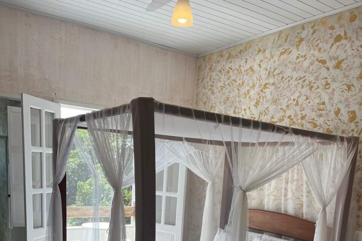 A room with a bed under a mosquito net, a wooden headboard, and a view of the outdoors.