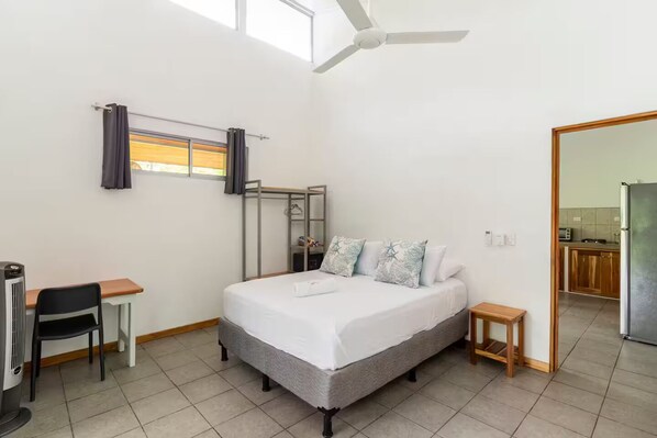 A bedroom with a bed, a desk, a chair, a small table, a ceiling fan, and a view of the kitchen.