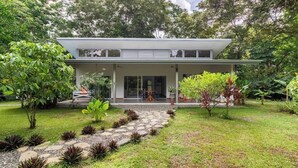 A modern two-story house with a covered patio, surrounded by lush greenery and a stone pathway leading to the entrance. - Complete house in the privacy of a spacious tropical setting (Puerto Jiménez)
