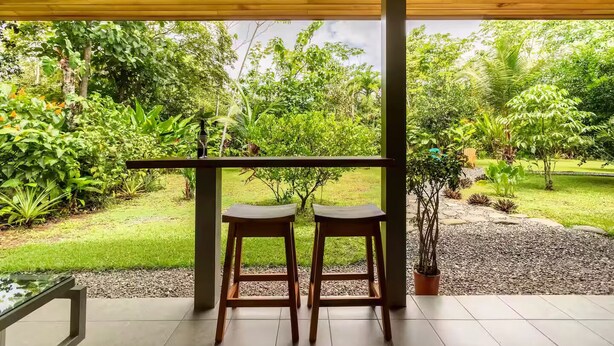 A covered outdoor seating area with two wooden stools, a glass table, and a view of a lush garden with various plants and trees. - Complete house in the privacy of a spacious tropical setting (Puerto Jiménez)