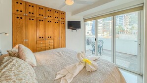 Un dormitorio con una cama grande, un armario de madera, un ventilador de techo, un televisor montado en la pared y un balcón con sillas.
