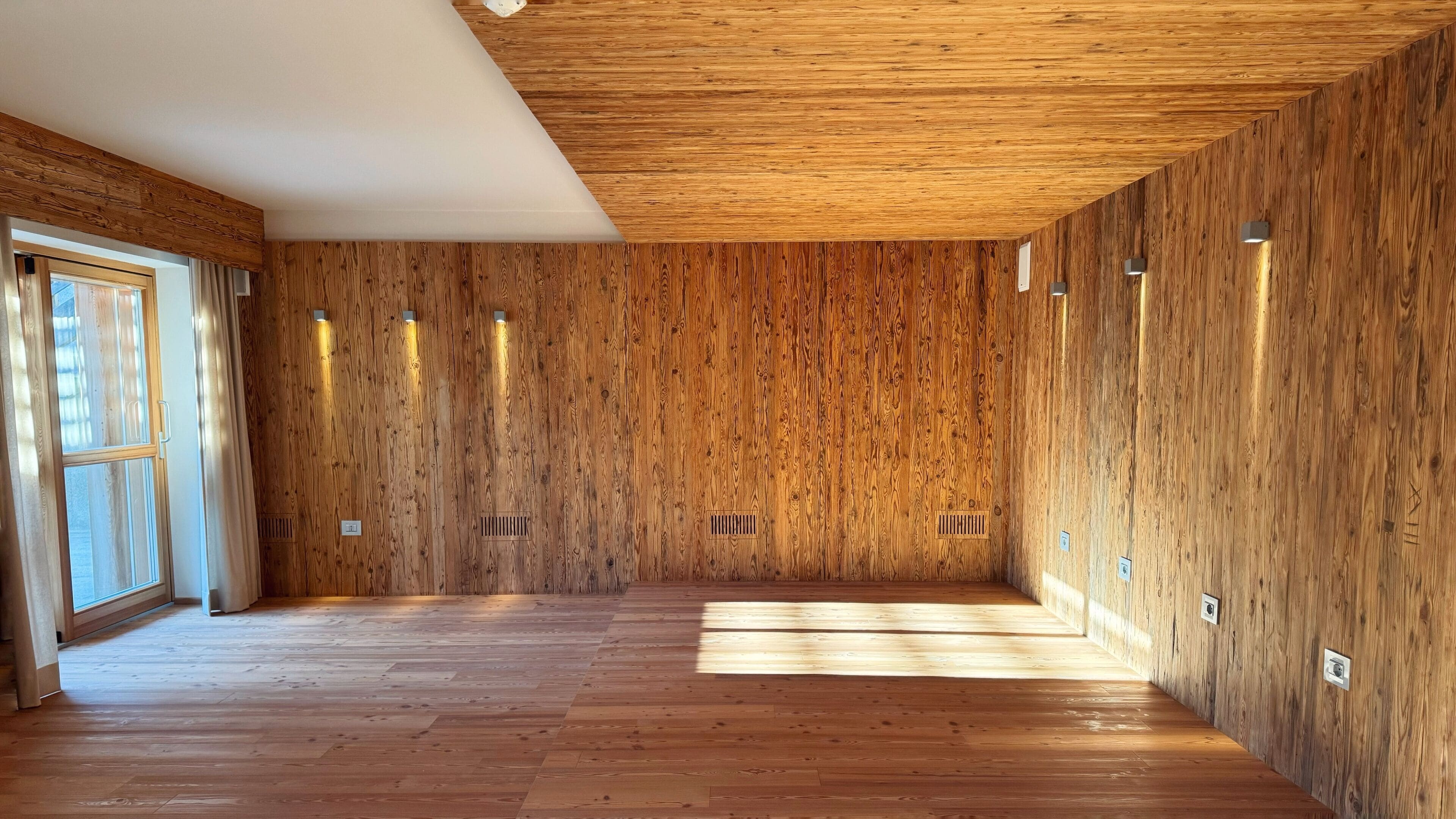 Ein geräumiger Raum mit Holzwänden und -böden, einem großen Fenster und integrierter Beleuchtung.