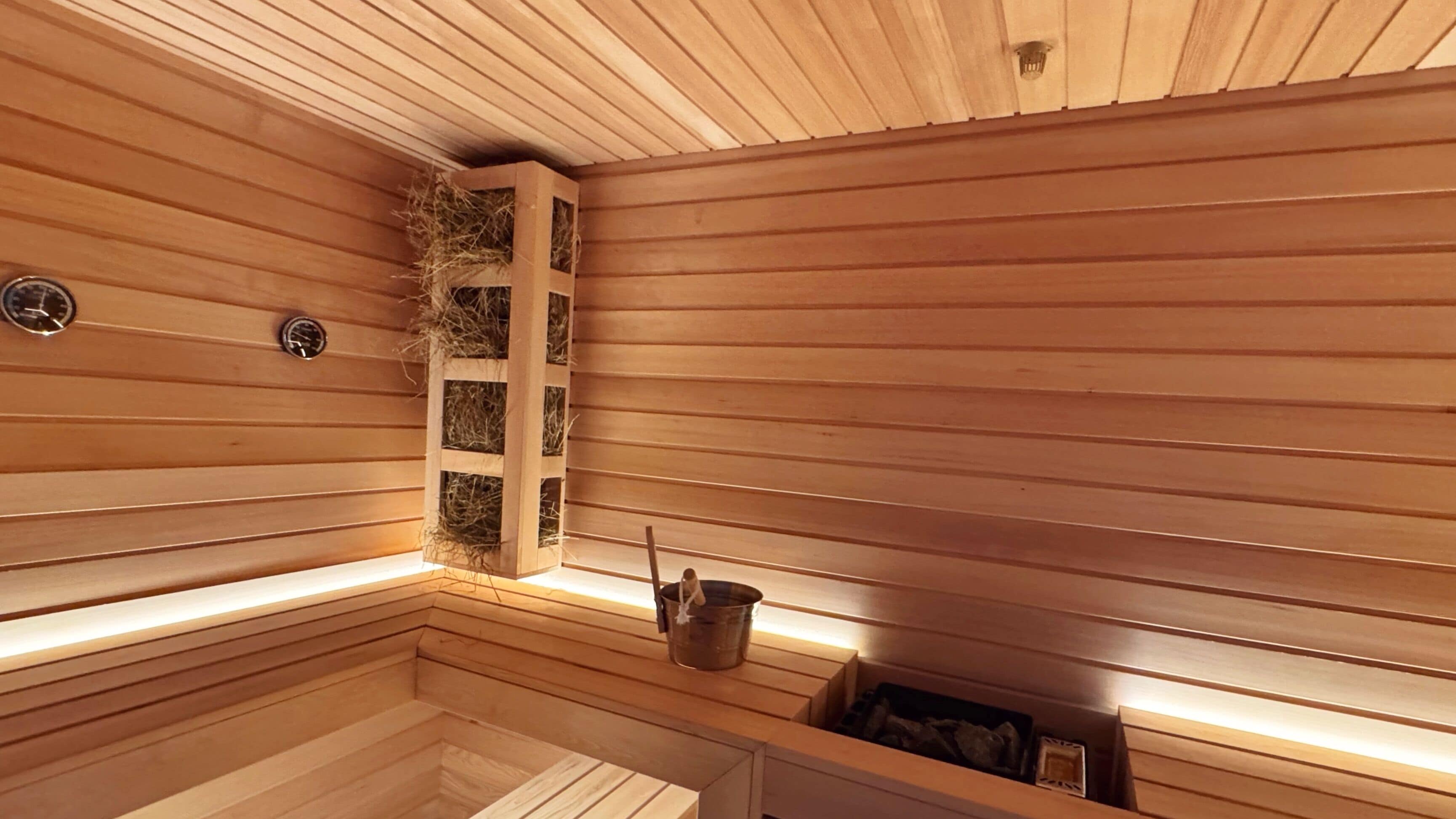 Un sauna aux murs et aux bancs en bois, une étagère en bois avec du foin et un seau contenant une serviette.