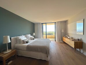 A modern bedroom with a large bed, a wooden dresser, and a view of the outdoors through a sliding glass door.