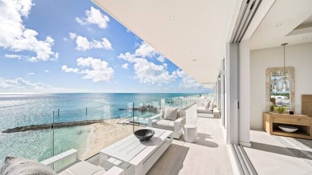 Un balcon moderne offrant une vue sur l’océan, avec des meubles blancs et une balustrade en verre.