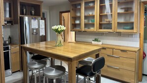 Una cocina moderna con armarios de madera, un refrigerador de acero inoxidable, una mesa de comedor de madera con taburetes tipo barra y un florero con flores blancas.