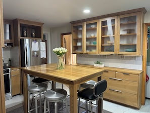 Una cocina moderna con armarios de madera, un refrigerador de acero inoxidable, una mesa de comedor de madera con taburetes tipo barra y un florero con flores blancas.