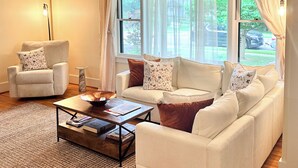 Una sala de estar con sofá, sillón, mesa de centro, ventanas grandes con cortinas y un tapete estampado.