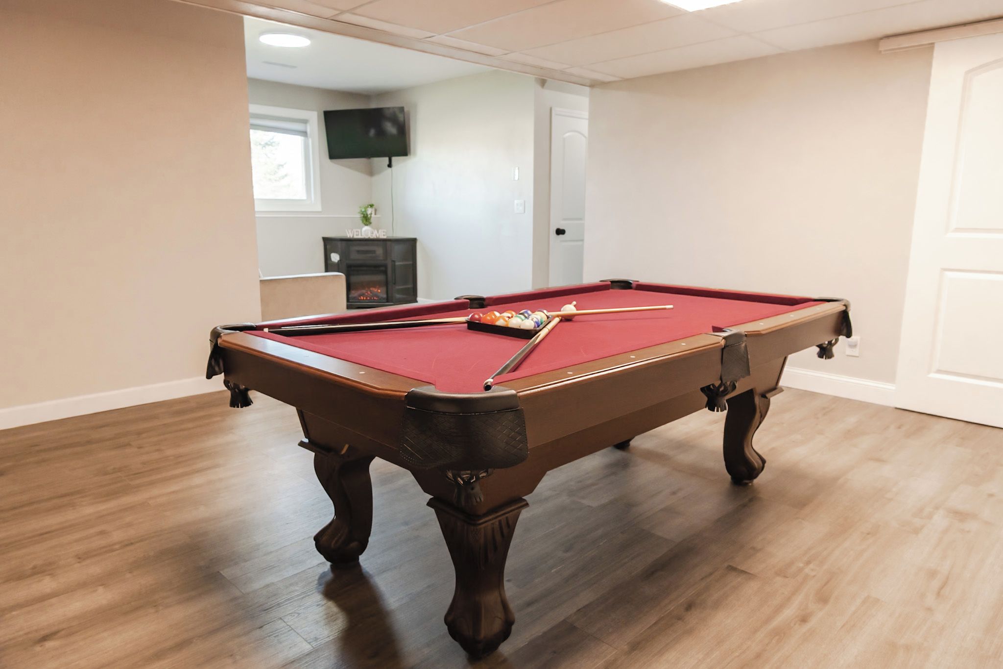 A pool table in a room with wooden flooring, a television on the wall, and a door.