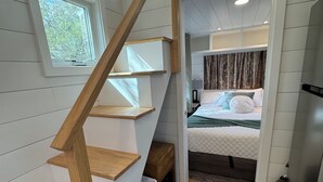 Une pièce de taille modeste avec un escalier en bois, un lit doté d’une tête de lit, une petite table et une poubelle.