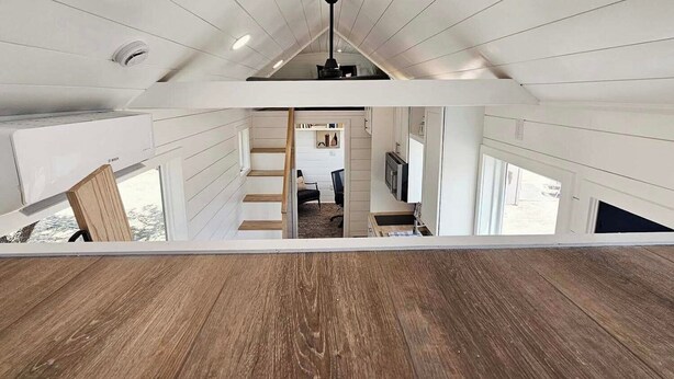 Une pièce aux hauts plafonds, dotée d’un plancher en bois, d’un escalier menant à un niveau supérieur et offrant une vue sur l’espace cuisine.