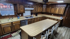 Une cuisine avec des meubles en bois, un îlot central doté de tabourets de bar, un ventilateur de plafond et une fenêtre donnant sur des maisons.