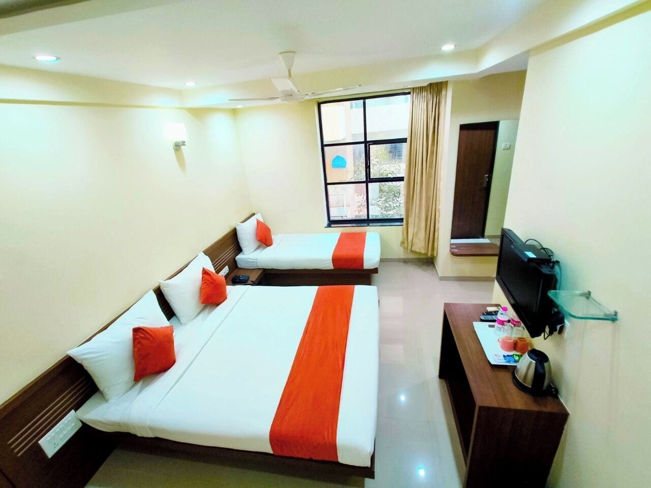A hotel room with two beds, a TV, a desk with a kettle, and a window with curtains.