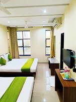 Ein Hotelzimmer mit zwei Betten, einem Fernseher, einem Schreibtisch mit Computer und einem fensterbepflanzten Fenster.
