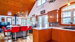 A modern kitchen with wooden cabinets, a bar area with red chairs, and large windows providing natural light. - Unforgettable Extended Stays In Beauce At The Lake (Saint-Victor)
