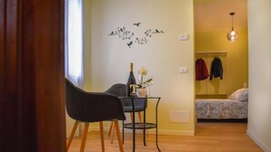 A small room with a wooden floor, a table with a bottle and flowers, a chair, and a bed with a floral bedspread. - B&B VILLA ELISA (Motta di Livenza)