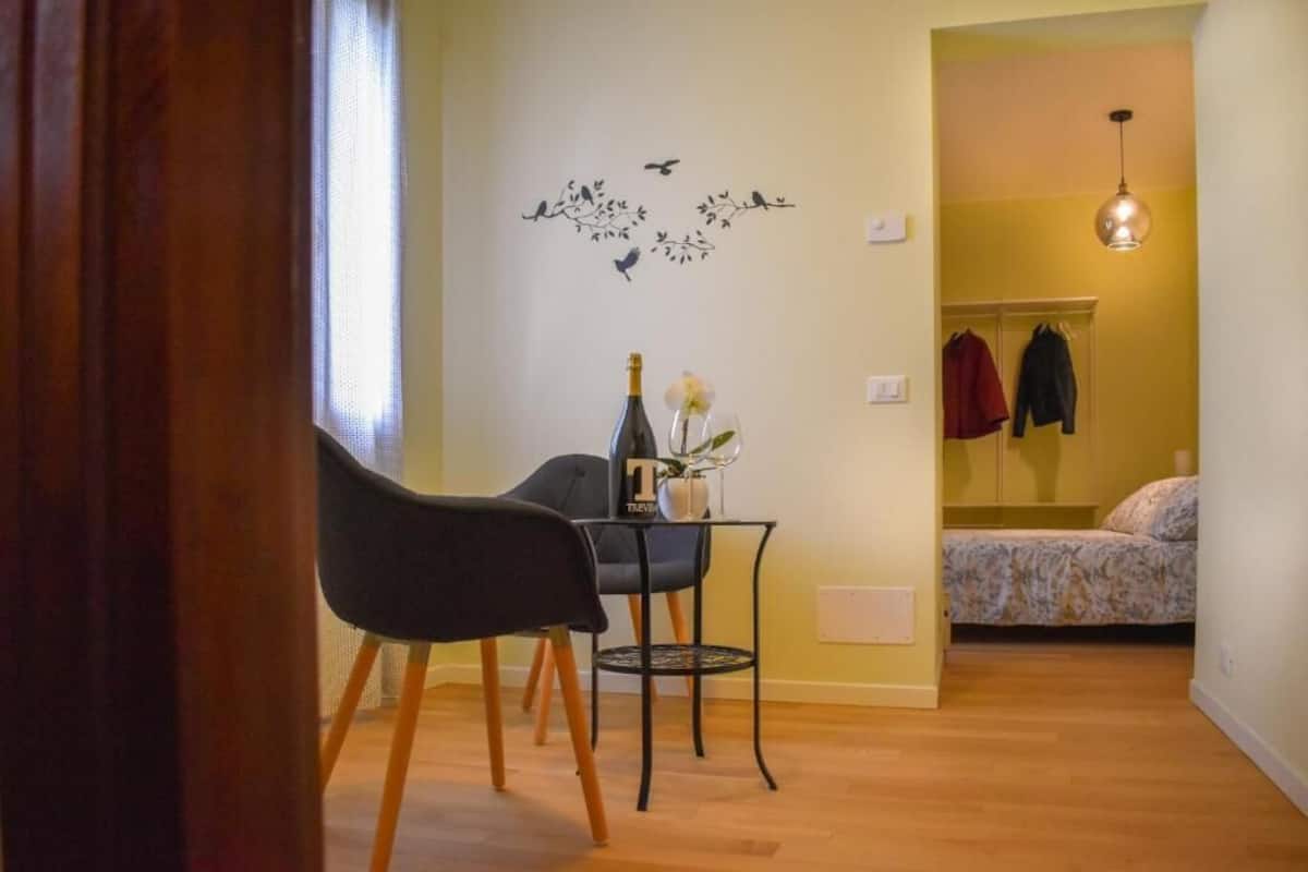 A small room with a wooden floor, a table with a bottle and flowers, a chair, and a bed with a floral bedspread.