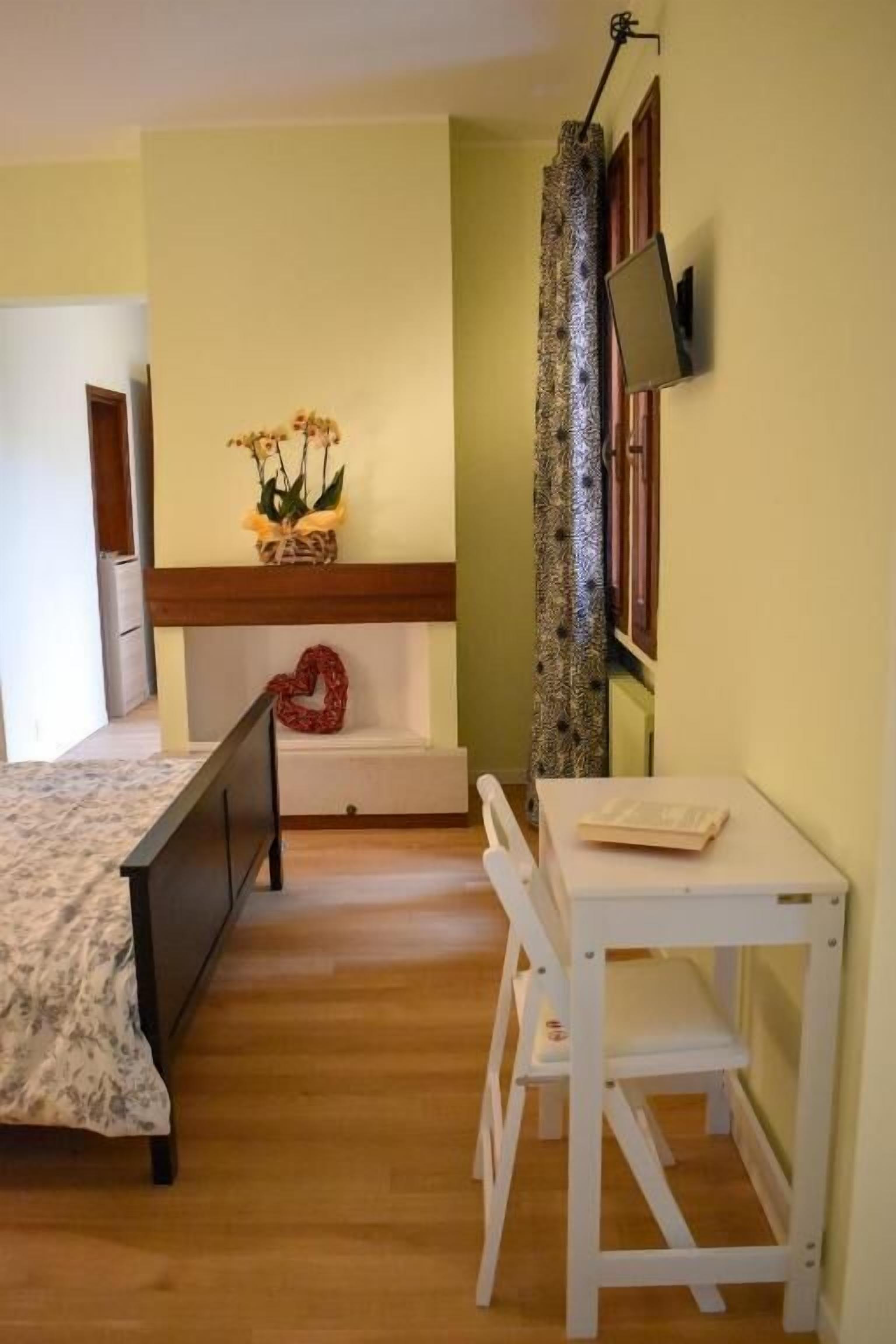 A bedroom with a bed, a small table, a chair, a TV mounted on the wall, and a decorative plant.