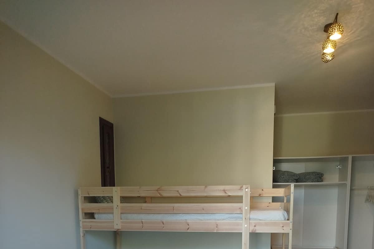 A bunk bed room with a wooden headboard, a lower bunk with a mattress and pillows, and a higher bunk with a mattress and pillows. There is a wardrobe with a mirror and a shelf above the bed.