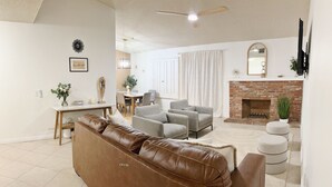 Un salon avec un canapé en cuir marron, des fauteuils gris, une table à manger avec des chaises, une cheminée et un ventilateur de plafond.