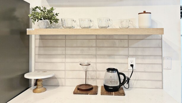 Une cuisine avec une étagère en bois où sont posées des tasses à café, une plante en pot et un récipient blanc. Sous l’étagère, on trouve un pare-feu blanc en carreaux, une petite table ronde avec un pied blanc et une bouilloire sur un plateau en bois.
