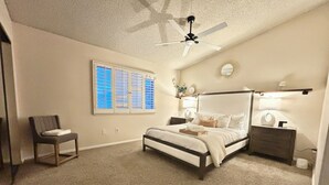 Une chambre à coucher comprenant un lit, une chaise, un ventilateur de plafond, une fenêtre avec des stores, une table de chevet avec une lampe et une commode avec un miroir.