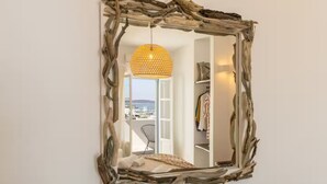 En spegel med en ram av naturligt trä som reflekterar ett rum med en hängande lampa, en stol och en utsikt över omgivningen.