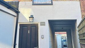 A narrow brick alleyway leading to a black door with a number plate, a window with a wheat pattern, and a small lantern on the roof.
