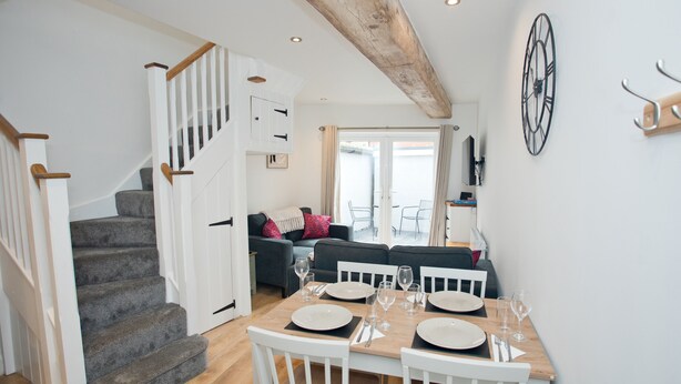 A dining area with a wooden table set for four, white chairs, and a staircase leading to a living area with a sofa and a TV.