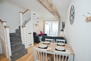 Un coin repas avec une table en bois dressée pour quatre personnes, des chaises blanches et un escalier menant à un espace de vie comprenant un canapé et un téléviseur.