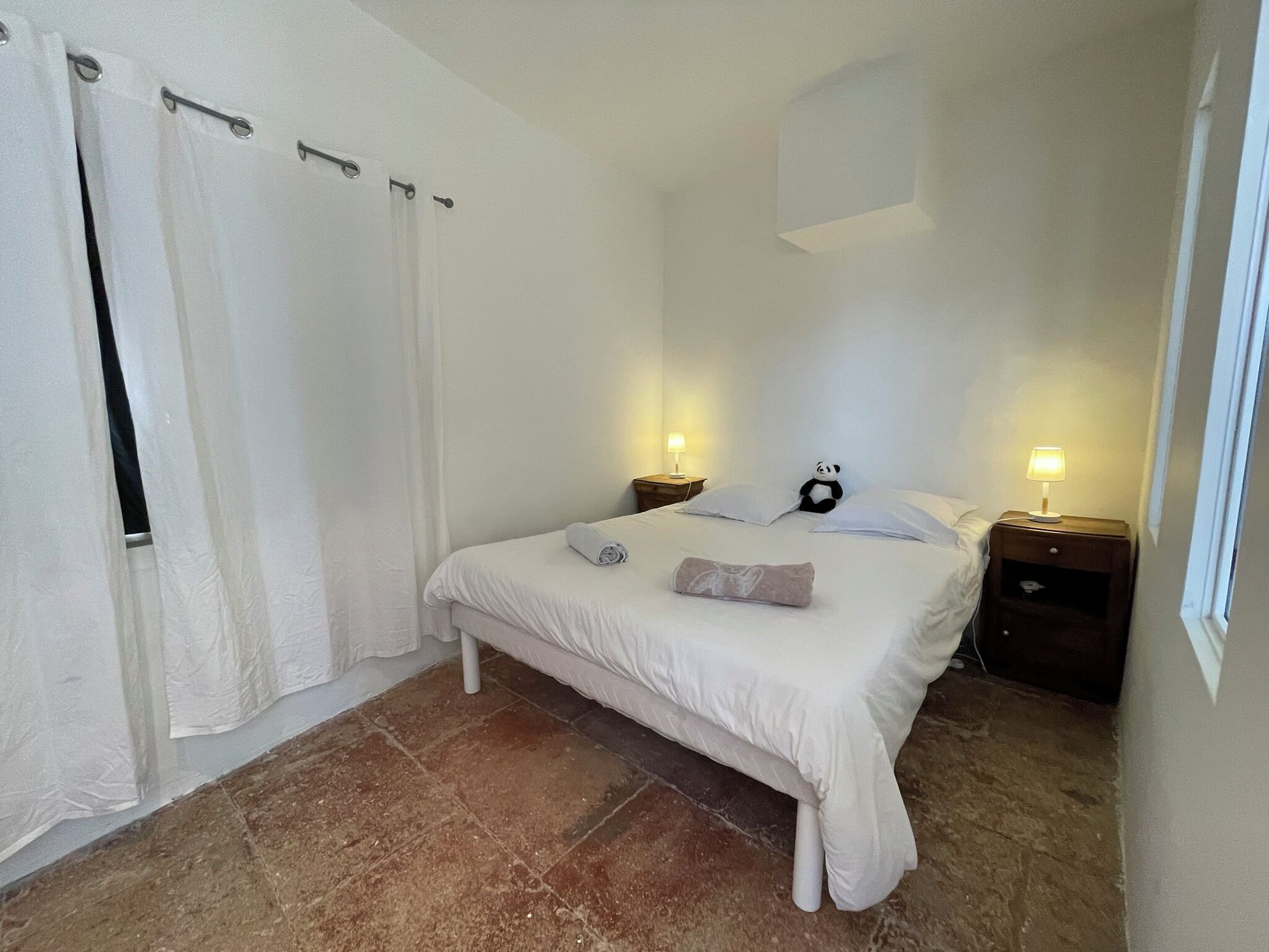 Ein Schlafzimmer mit einem Bett, zwei Nachttischen mit Lampen, einem Fenster und einem Vorhang.