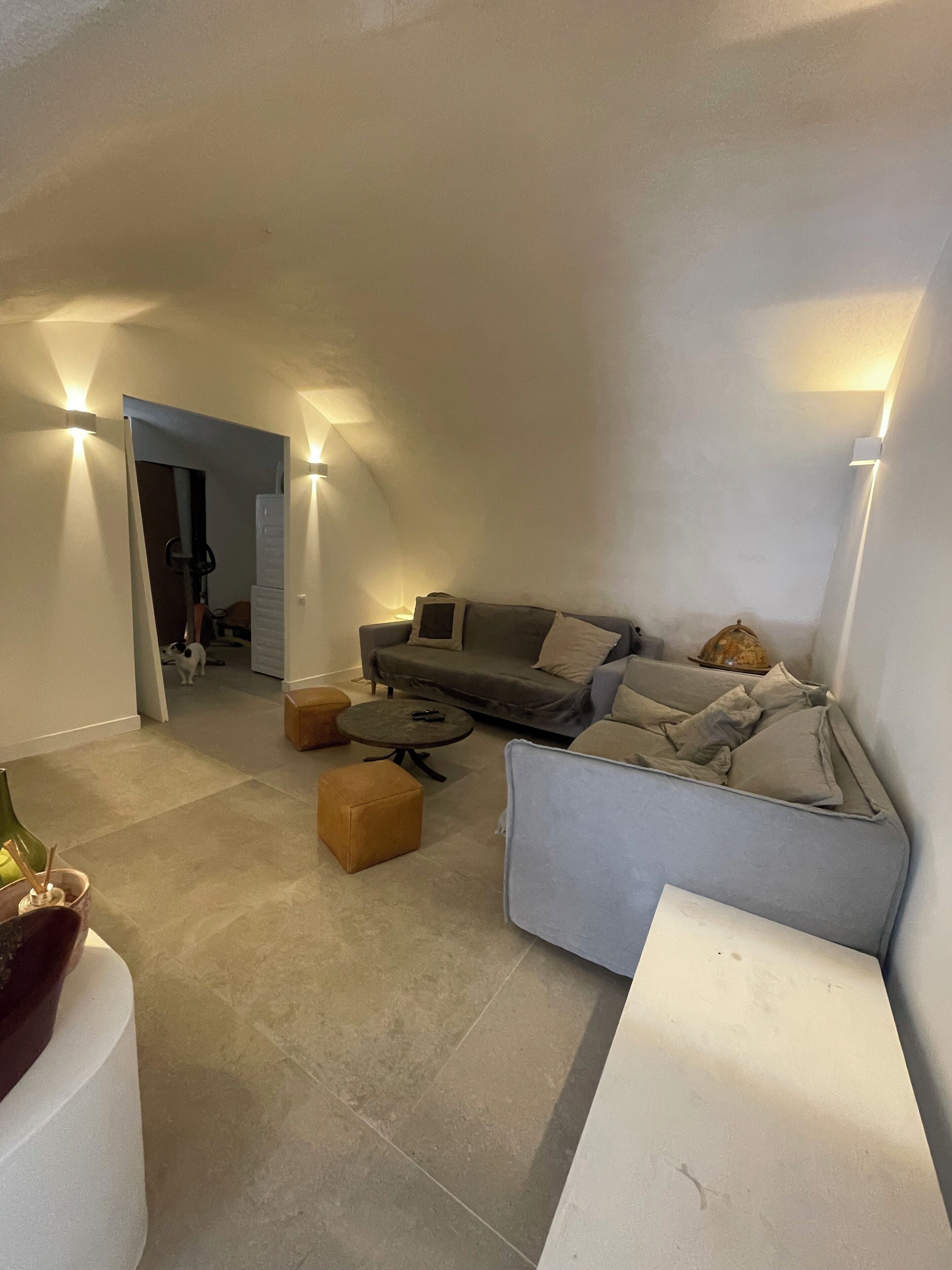 A modern living room with a grey sofa, two ottomans, a coffee table, and a white side table. There are wall-mounted lights and a door leading to another room.